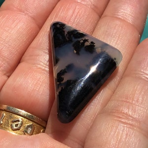 May include: A triangular, translucent, grey and black agate gemstone with dendritic patterns. The gemstone is approximately 5 cm in length.