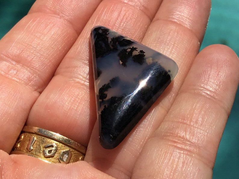 May include: A triangular, translucent, grey and black agate gemstone with dendritic patterns. The gemstone is approximately 5 cm in length.