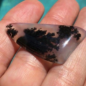 May include: A translucent, light purple agate cabochon with black dendritic inclusions. The agate is shaped like a triangle and has a smooth, polished surface.