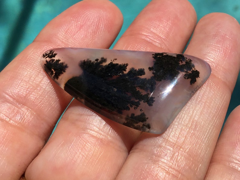 May include: A translucent, light purple agate cabochon with black dendritic inclusions. The agate is shaped like a triangle and has a smooth, polished surface.