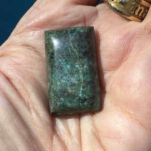 May include: A dark green, rectangular, polished gemstone with a marbled pattern. The stone is smooth and has a slight sheen.