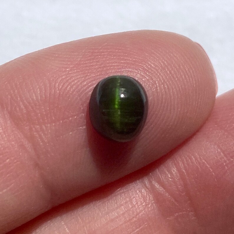 May include: A dark green oval cabochon gemstone with a chatoyant effect. The stone has a smooth, polished surface and a dark green color with a hint of black.