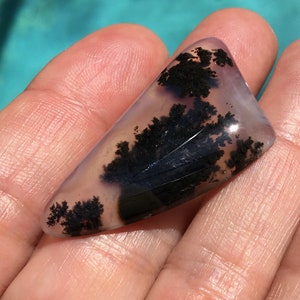 May include: A translucent, light purple agate cabochon with black dendritic inclusions. The agate has a smooth, polished surface and is irregularly shaped.