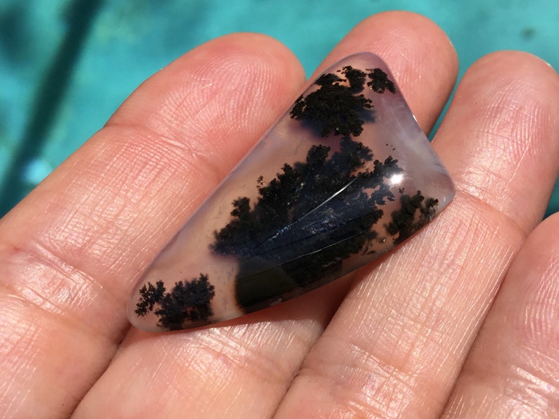 May include: A translucent, light purple agate cabochon with black dendritic inclusions. The agate has a smooth, polished surface and is irregularly shaped.