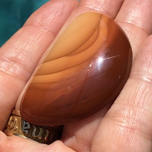 Pode incluir: Uma pedra de &aacute;gata marrom lisa e polida com faixas laranja e brancas. A pedra tem a forma de uma l&aacute;grima e um acabamento brilhante.