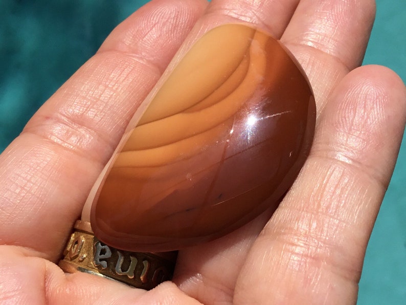 Pode incluir: Uma pedra de &aacute;gata marrom lisa e polida com faixas laranja e brancas. A pedra tem a forma de uma l&aacute;grima e um acabamento brilhante.