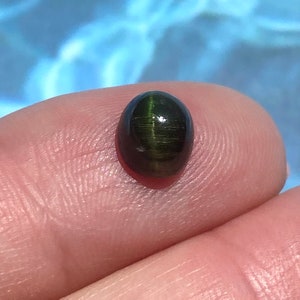 May include: A dark green oval cabochon gemstone with a chatoyant effect. The stone is set in a bezel setting and has a high polish.