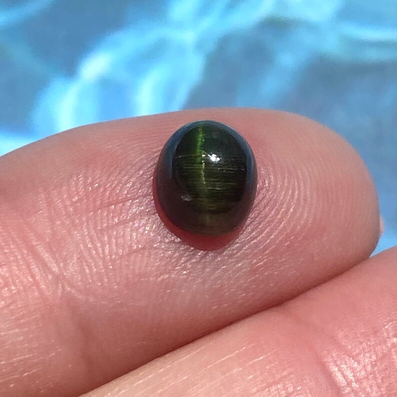 May include: A dark green oval cabochon gemstone with a chatoyant effect. The stone is set in a bezel setting and has a high polish.