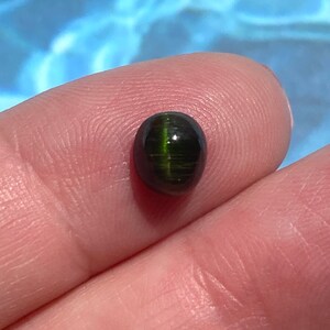May include: A dark green oval cabochon gemstone with a shimmering, chatoyant effect. The stone has a smooth, polished surface and is set in a bezel setting.