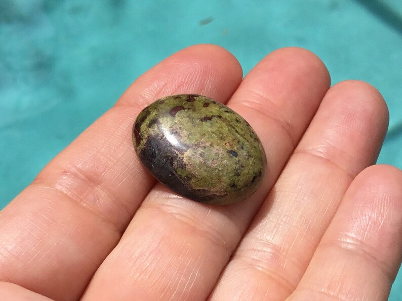 May include: A green and brown oval-shaped gemstone with red and black flecks. The gemstone is polished and smooth.
