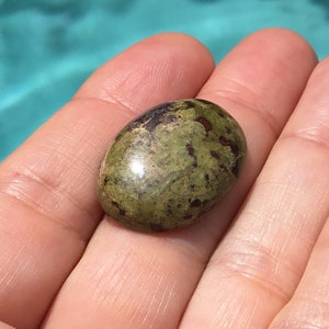 May include: A green and brown oval-shaped gemstone with a smooth, polished surface. The stone has a mottled pattern of dark green and brown.