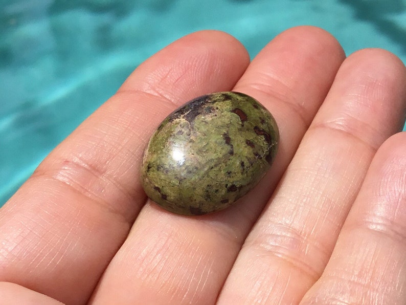 May include: A green and brown oval-shaped gemstone with a smooth, polished surface. The stone has a mottled pattern of dark green and brown.
