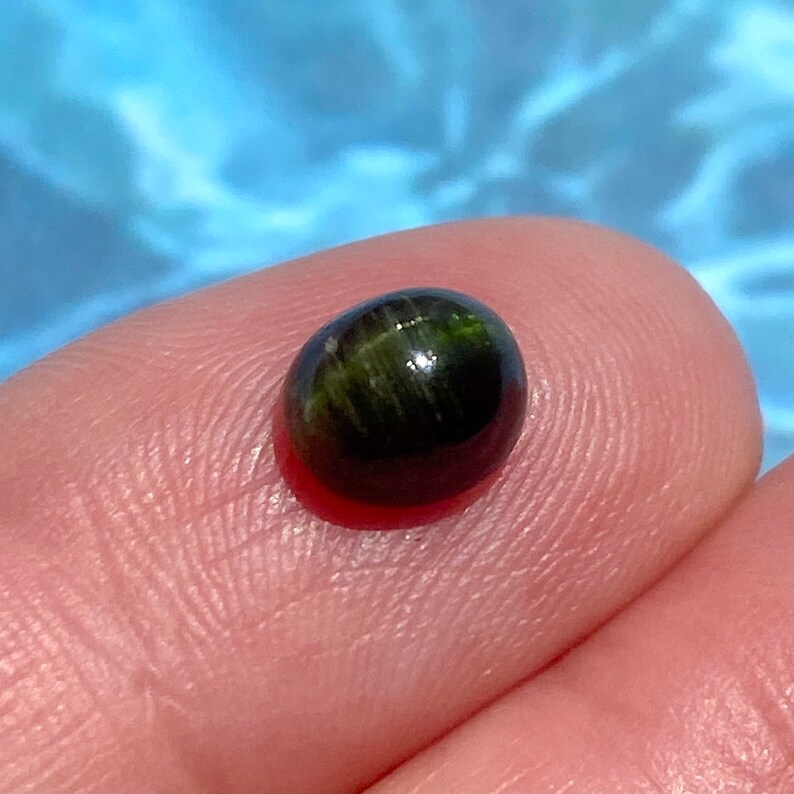 May include: A dark green oval cabochon gemstone with a chatoyant effect. The stone has a black background with green and gold streaks.