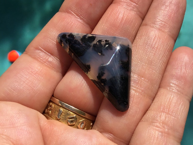 May include: A triangular, translucent, grey and black agate cabochon with dendritic inclusions. The cabochon is approximately 5 cm in length.