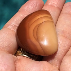 Pode incluir: Uma pedra de &aacute;gata lisa e polida, de cor castanho claro, com um padr&atilde;o de redemoinho de tons castanhos escuros e laranja.