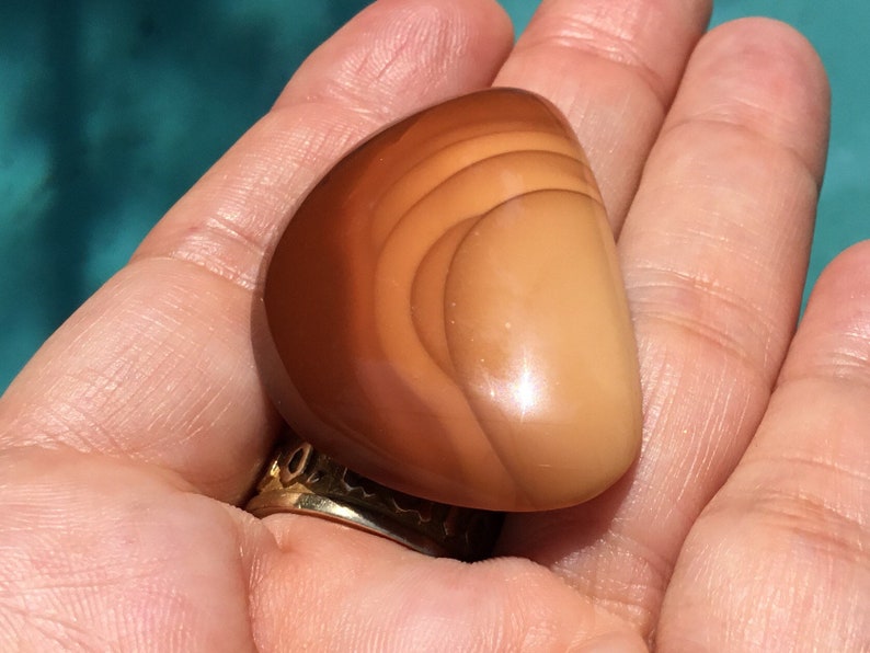 Pode incluir: Uma pedra de &aacute;gata lisa e polida, de cor castanho claro, com um padr&atilde;o de redemoinho de tons castanhos escuros e laranja.