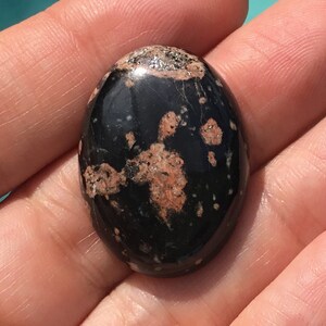 May include: A black and pink oval cabochon gemstone with a speckled pattern. The gemstone is polished and smooth.