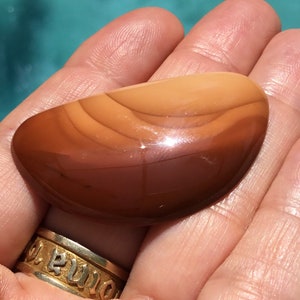 Pode incluir: Uma pedra de &aacute;gata lisa e polida, de cor marrom e laranja, com um padr&atilde;o em camadas. A pedra tem formato oval e um acabamento brilhante.