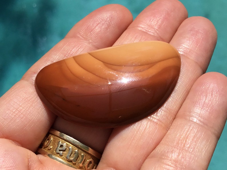 Pode incluir: Uma pedra de &aacute;gata lisa e polida, de cor marrom e laranja, com um padr&atilde;o em camadas. A pedra tem formato oval e um acabamento brilhante.