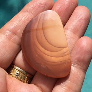 Pode incluir: Uma pedra de &aacute;gata lisa de cor castanho claro com an&eacute;is conc&ecirc;ntricos de castanho escuro. A pedra tem a forma de uma l&aacute;grima e tem uma superf&iacute;cie polida.
