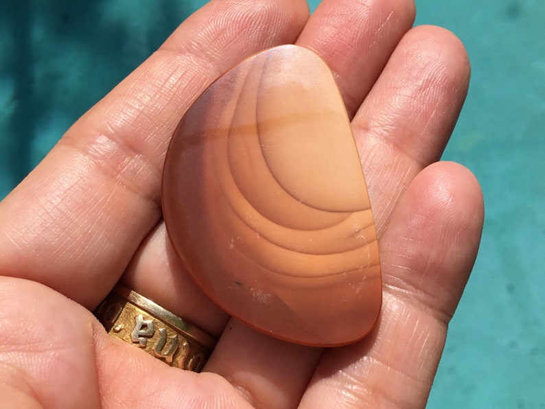 Pode incluir: Uma pedra de &aacute;gata lisa de cor castanho claro com an&eacute;is conc&ecirc;ntricos de castanho escuro. A pedra tem a forma de uma l&aacute;grima e tem uma superf&iacute;cie polida.