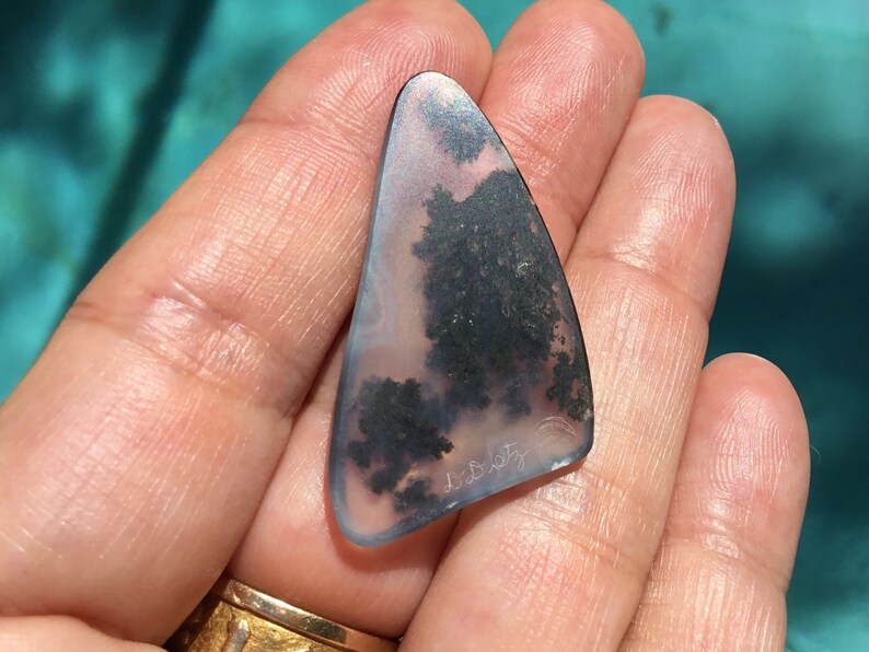 May include: A triangular piece of agate with a dendritic pattern in shades of black, grey, and white. The agate is translucent and has a slight pink hue. The piece is approximately 5 cm long and 4 cm wide. The agate is signed "D. Beatty".
