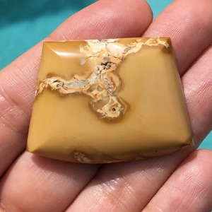 May include: A light brown, square-shaped cabochon gemstone with a unique pattern of white and brown veins. The stone has a smooth, polished surface.