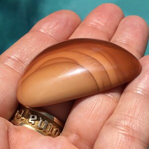 Pode incluir: Uma pedra de &aacute;gata lisa e polida, de cor castanho claro, com um padr&atilde;o de bandas sobrepostas. A pedra tem forma oval e um acabamento brilhante.