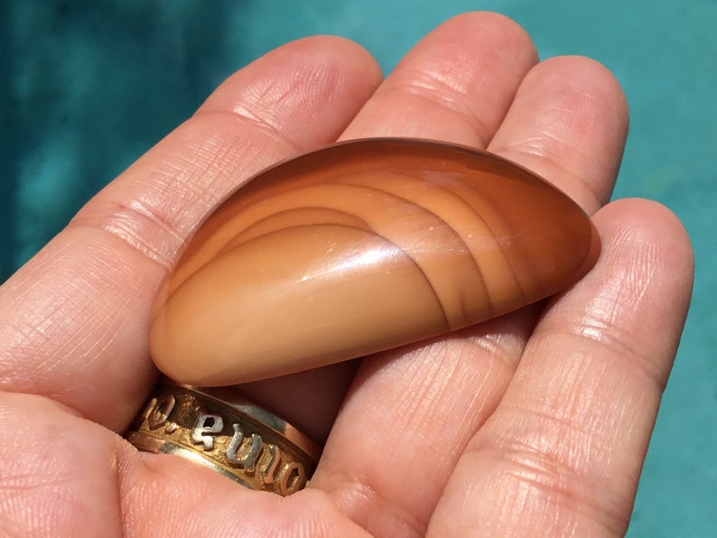 Pode incluir: Uma pedra de &aacute;gata lisa e polida, de cor castanho claro, com um padr&atilde;o de bandas sobrepostas. A pedra tem forma oval e um acabamento brilhante.