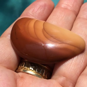 Pode incluir: Uma pedra de &aacute;gata lisa e polida, de cor marrom e laranja, com um padr&atilde;o em camadas. A pedra tem a forma de uma meia lua.