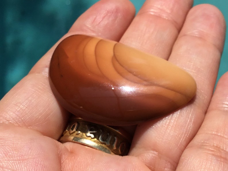 Pode incluir: Uma pedra de &aacute;gata lisa e polida, de cor marrom e laranja, com um padr&atilde;o em camadas. A pedra tem a forma de uma meia lua.