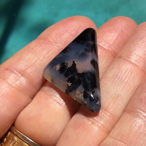 May include: A triangular shaped, polished agate cabochon with a dendritic pattern in black and white against a light blue and pink background.