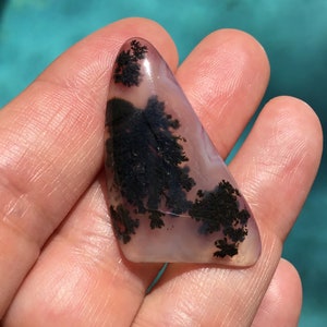 May include: A triangular shaped, polished agate stone with dendritic inclusions. The stone is translucent and has a light pink and white base with black dendritic patterns.