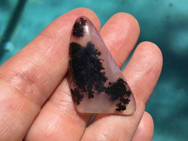 May include: A triangular shaped, polished agate stone with dendritic inclusions. The stone is translucent and has a light pink and white base with black dendritic patterns.