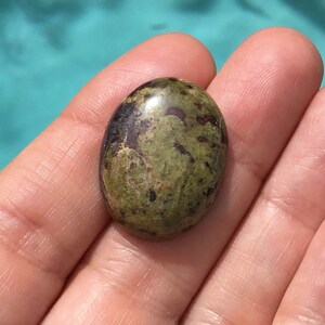 May include: A green and brown oval cabochon gemstone with a polished surface. The stone has a mottled pattern of green, brown, and black.