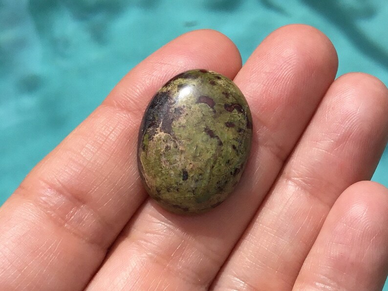 May include: A green and brown oval cabochon gemstone with a polished surface. The stone has a mottled pattern of green, brown, and black.