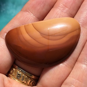 Pode incluir: Uma pedra de &aacute;gata lisa e polida, de cor marrom e laranja, com bandas conc&ecirc;ntricas de cor. A pedra tem formato oval e um acabamento brilhante.
