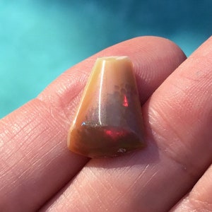 May include: A small, triangular, iridescent opal cabochon with a brown, red, and white color pattern.