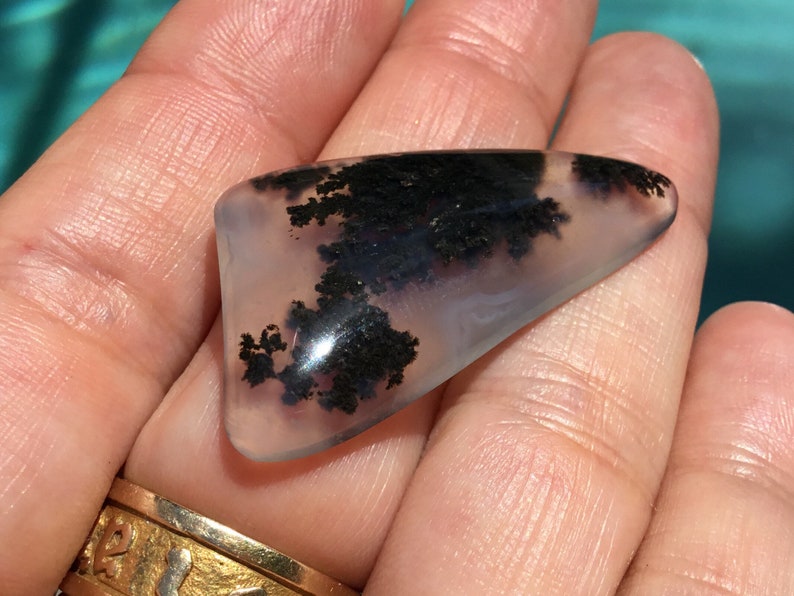 May include: A translucent, light grey agate cabochon with black dendritic inclusions. The cabochon is shaped like a triangle with rounded edges.