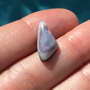 May include: A small, triangular, iridescent opal gemstone with a black backing. The opal has a white base with flashes of rainbow colors.