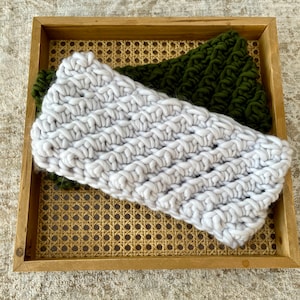 May include: Two knitted headbands, one white and one dark green, are displayed in a woven basket. The white headband has a chunky knit texture.