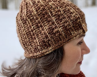Op de afbeelding: Een gebreide beanie in bruin en beige met een leren patch aan de zijkant. De beanie wordt gedragen door een persoon met bruin haar.