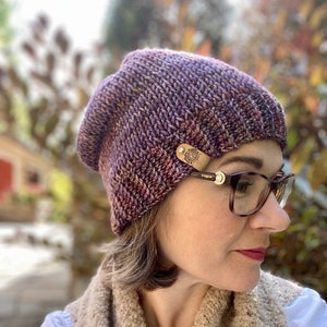 May include: A purple and brown knit beanie with a leather patch that says "Rogue" on it. The beanie is being worn by a person with brown hair and glasses.