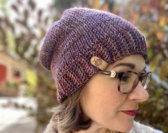 Könnte beinhalten: Eine lila und braune Strickmütze mit einem Lederpatch, auf dem "Rogue" steht. Die Mütze wird von einer Person mit braunen Haaren und Brille getragen.