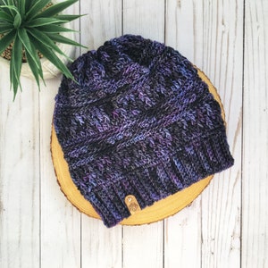May include: A purple and black knitted beanie with a brown leather tag. The beanie is sitting on a wooden surface.