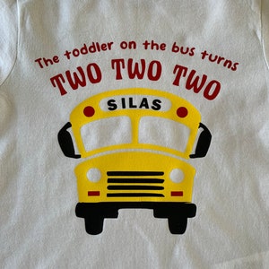 Puede incluir: Camiseta blanca para niño con un diseño de autobús escolar amarillo. El texto rojo dice "The toddler on the bus turns TWO TWO TWO", con "SILAS" en el autobús.