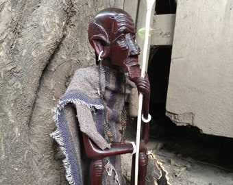 May include: A wooden sculpture of a seated figure wearing a patterned cloth. The figure holds a long, thin staff in its right hand. The sculpture is carved in a dark brown colour and has a detailed face.