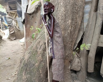 May include: A wooden figurine of a person standing with a staff. The figurine is carved in a dark brown color and is wearing a long, flowing robe. The figurine is standing on a wooden base.