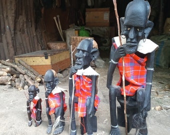 May include: Five wooden figurines of people wearing red and black patterned clothing. The figurines are carved in a traditional style and are standing on a wooden base.