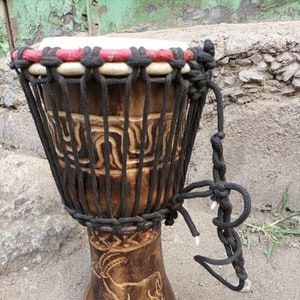 May include: A brown wooden djembe drum with a black rope handle and a carved animal design. The drum has a white head with a red and black trim.